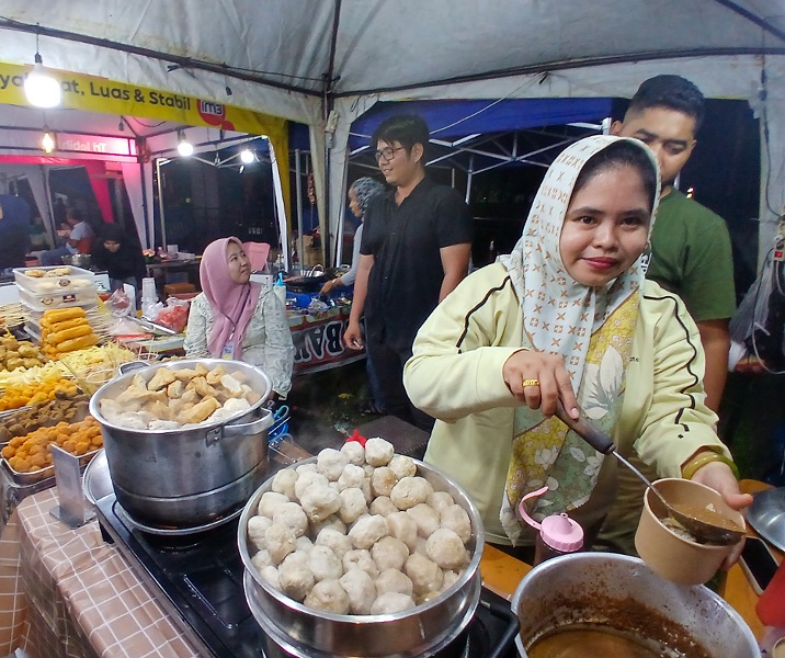 Ical berdiri mendampingi istrinya saat menyiapkan menu bakso bagi pembeli di area Gelanggan Remaja Pekanbaru. Foto: Surya/Riau1.
